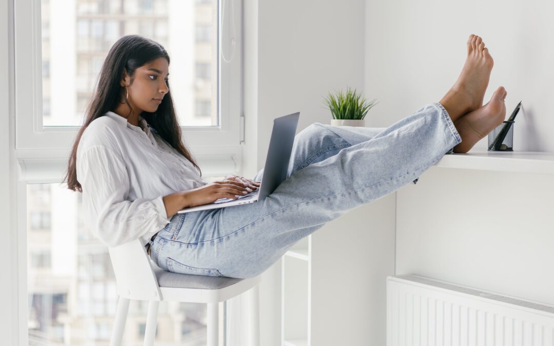 Young barefoot woman with long dark hair wearing a long-sleeved white blouse and faded jeans, seated with her feet up in an all-white room