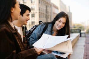 Two female and one male students standing on a college campus, wearing light jackets and holding papers