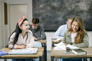 Four young adults, two female and two male, at desks in a classroom with a blackboard behind them