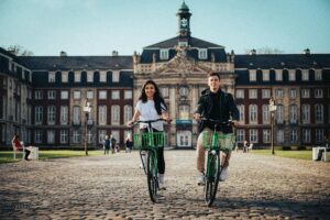 One male and one female student on a college campus on green bicycles headed towards the viewer