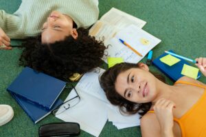 Two dark-haired teen students lying on the floor with books, pencils and notebooks.