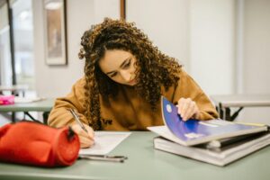 Female teen with long brown hair and long-sleeved brown shirt at school desk with pen, paper and notebooks