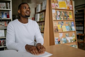 Young adult black male student wearing white long-sleeved T-shirt reading Braille in library